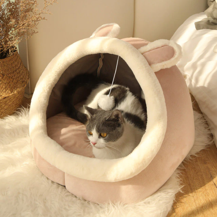 Beige cat bed with cream rim and dangling pom-pom toy; enclosed cozy sleeping nest