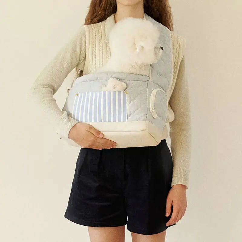 Cream pet sling carrier with striped front pocket and soft hood, worn on shoulders