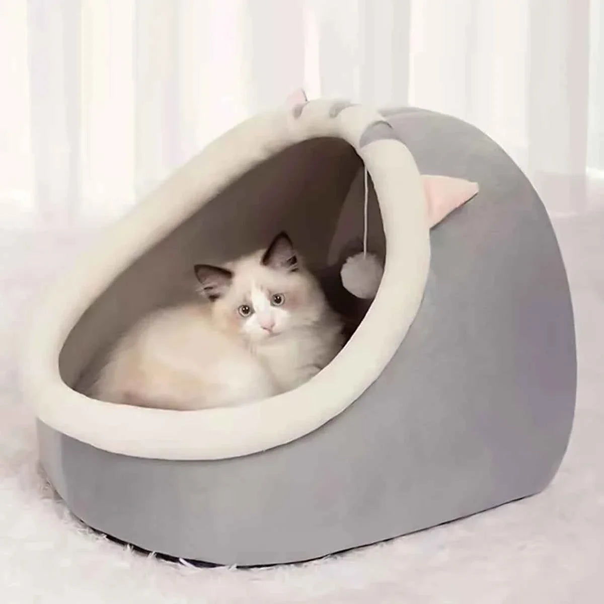 Grey semi-enclosed pet bed with cream inner rim and hanging pom; white British Shorthair