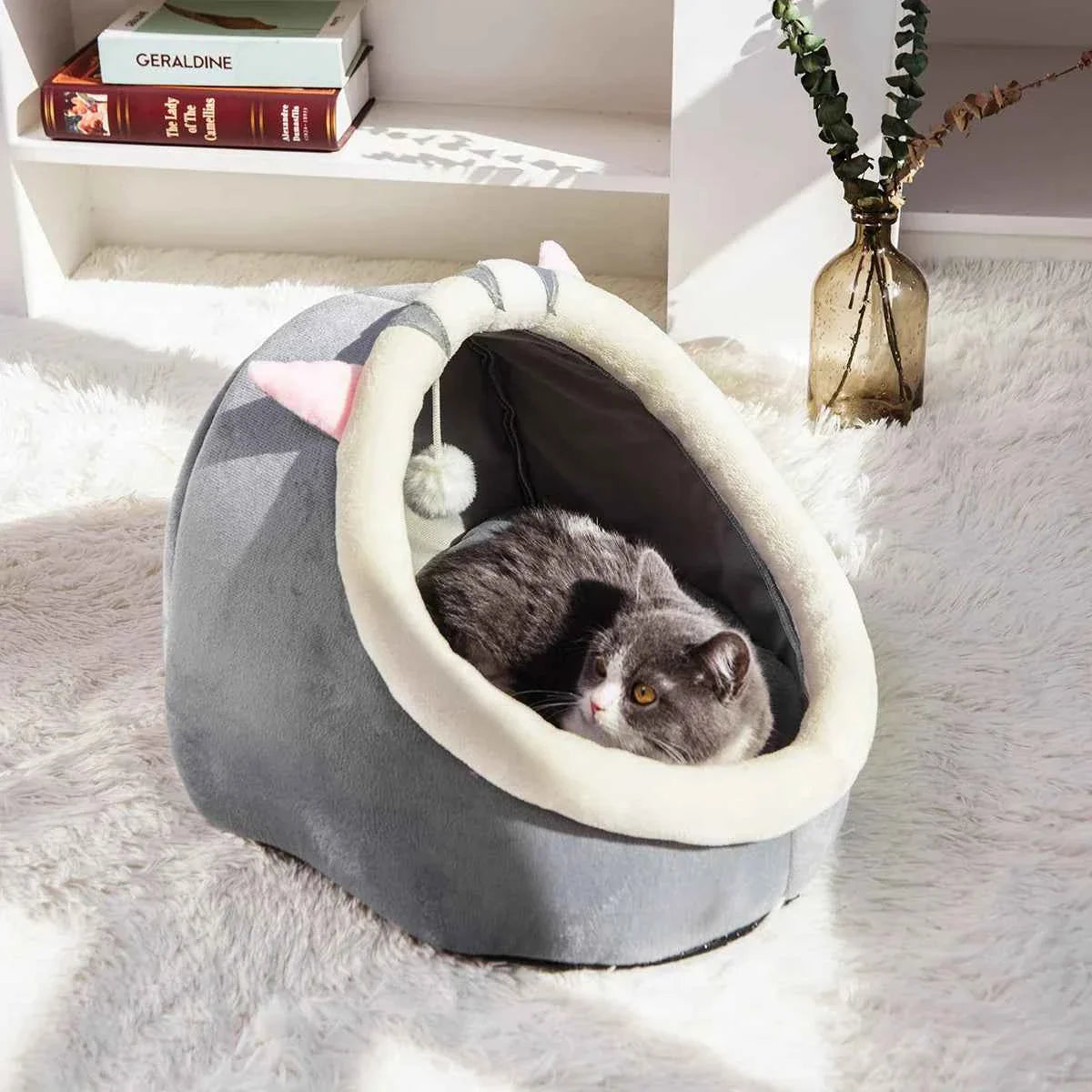 Grey semi-enclosed cat bed with cream rim and hanging pom; British Shorthair curled up